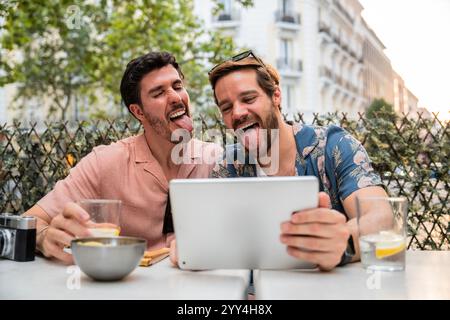 A joyful gay couple sits at an outdoor cafe, playfully sticking their tongues out while using a digital tablet, showcasing a moment of fun and connect Stock Photo