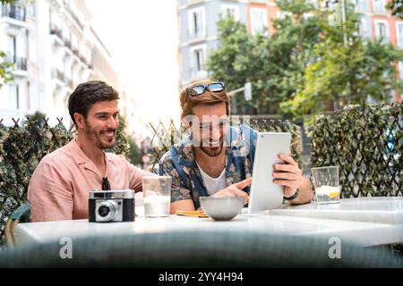 Two men joyfully share a moment using a tablet at an outdoor cafe, embodying casual, modern LGBTQ+ life Stock Photo