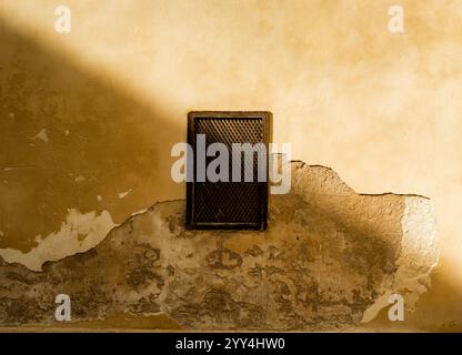 Textured old wall with peeling plaster and a small diamond-patterned window illuminated by soft sunlight and shadow Stock Photo