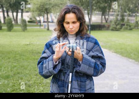 A blind woman stands in a park, using a smartphone with her left hand while holding a white cane in her right She wears a blue plaid coat, surrounded Stock Photo