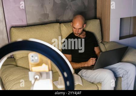 A man sits on a couch with a laptop, focused on social media content creation A smartphone and ring light setup are visible, indicating a casual but p Stock Photo
