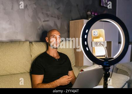 A man sitting on a couch creating social media content He is using a smartphone mounted on a ring light, highlighting modern media work at home Stock Photo