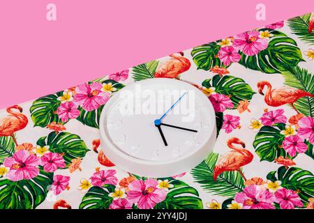 A minimalistic white wall clock lies on a vibrant tropical fabric adorned with flamingos, hibiscus flowers, and palm leaves. The colorful backdrop cre Stock Photo