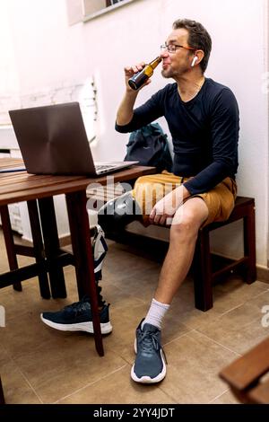 A man with a prosthetic leg sits at an outdoor table, using a laptop and drinking from a bottle He wears headphones, a dark shirt, and shorts, enjoyin Stock Photo