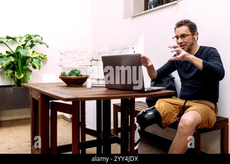 A man wearing a prosthetic leg sits at an outdoor table, actively participating in a video call on his laptop The setting includes lush plants and a c Stock Photo