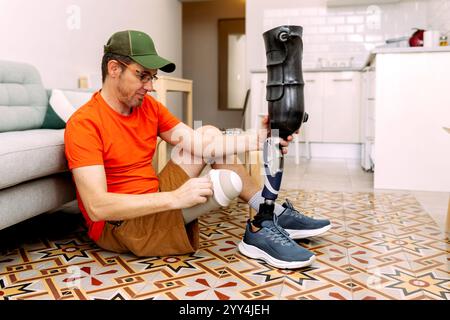 A man sitting on a tiled floor in a cozy home setting, assembling his prosthetic leg He is wearing a green cap and an orange shirt, focusing on adjust Stock Photo