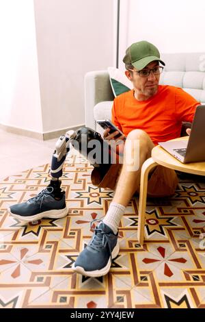 A man with a prosthetic leg sits on a vibrant patterned floor using a laptop and smartphone He wears casual attire, blending technology with everyday Stock Photo