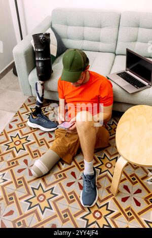 A man with a prosthetic leg sits on a patterned floor, focused on his smartphone A laptop rests on the nearby couch, capturing a modern, adaptive life Stock Photo