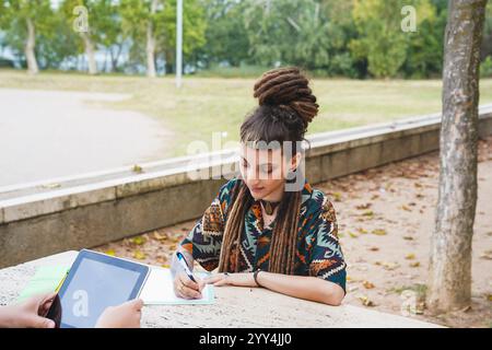 An agender person, distinguishable by their dreadlocks and fringe, engages in writing near an unrecognizable person with tablet Stock Photo