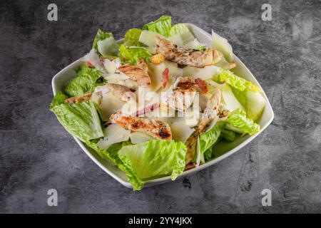 A fresh Caesar salad featuring tender grilled chicken, crisp romaine lettuce, shaved Parmesan cheese, bacon, and croutons, all in a white bowl on a gr Stock Photo