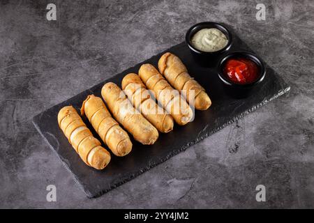A slate board holds six golden, crispy tequeños, accompanied by two savory dipping sauces. Perfect for appetizers or snacks, offering a delightful cul Stock Photo