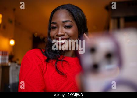 Young black woman wearing red sweater taking selfie with smartphone in warm colored cafe Stock Photo