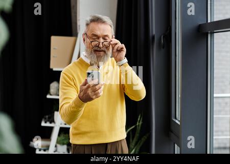 Mature man in a yellow sweater using his smartphone while standing in a stylish room Stock Photo