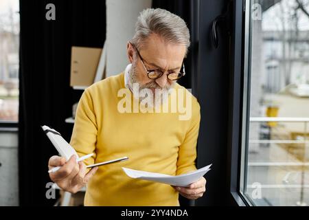 Mature man in yellow sweater reviews important papers while standing near a window. Stock Photo
