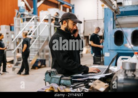 Male warehouse worker talking on smart phone while using laptop in manufacturing factory Stock Photo
