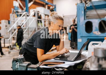 Female warehouse worker talking on smart phone while using laptop in manufacturing factory Stock Photo
