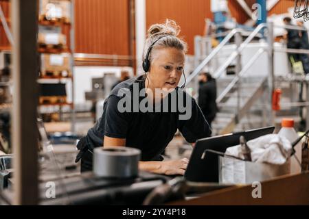 Female warehouse worker wearing headset while using laptop in manufacturing factory Stock Photo