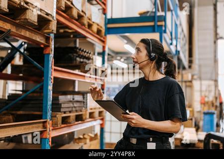Female worker wearing headset while taking inventory and holding tablet PC in distribution warehouse Stock Photo