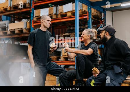 Male and female warehouse workers eating burgers while talking with each other in factory Stock Photo