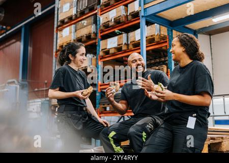 Happy male and female warehouse workers eating burgers while enjoying and talking with each other in factory Stock Photo