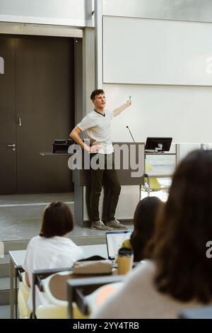 Male professor standing with hand on hip while teaching students in lecture hall at university Stock Photo