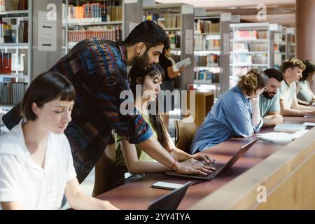 Man assisting female friend using laptop at desk in college library Stock Photo
