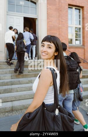Portrait of smiling woman carrying bag while looking over shoulder at college campus Stock Photo