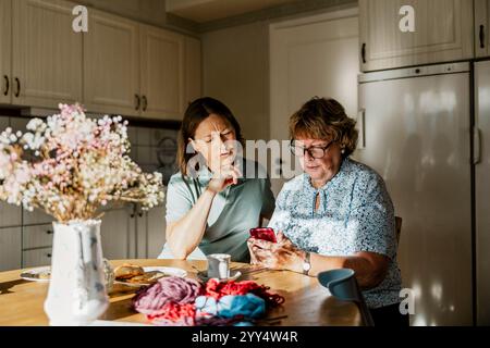 Female nurse with hand on chin sitting next to senior woman using smart phone at home Stock Photo