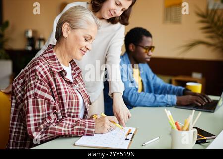 Side view of smiling mature female employee writing in calendar table on clipboard while colleague pointing at schedule planning together monthly tasks at working desk in office, copy space Stock Photo