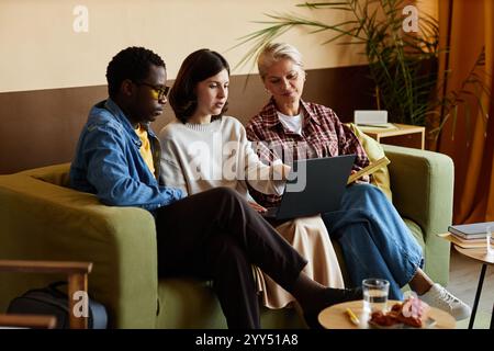 Multigenerational team of office workers discussing project details pointing at laptop screen while sitting on couch at workplace Stock Photo
