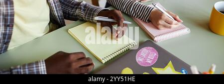 Website header shot of two colleagues from different ethnic backgrounds writing in colorful notebooks collaborating at working desk in office Stock Photo
