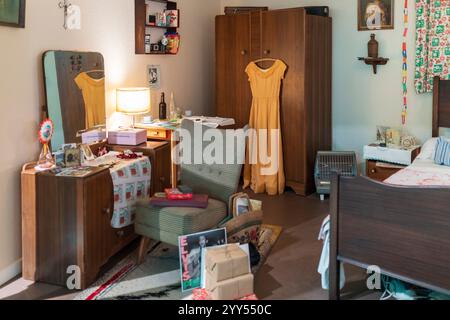 London, UK, December 4, 2024: Room illustrating the interior of an English house from the 1960s, Museum of the Home. A dress hangs on a wardrobe, chai Stock Photo