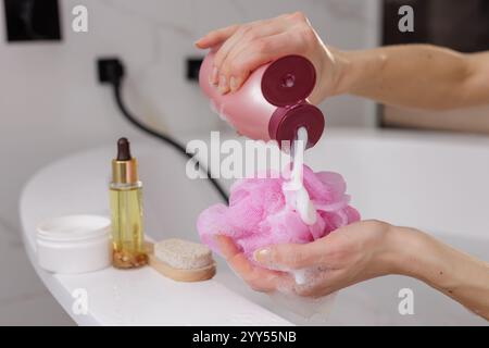 woman's hands applying shower gel on pink washcloth against bathroom background, body and hair care products lying nearby Stock Photo