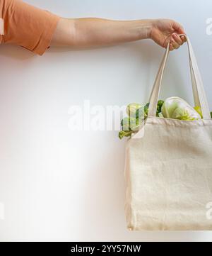 A hand holding a cotton bag filled with vegetables and greens against a white background, symbolizing an eco-friendly lifestyle and the rejection of Stock Photo