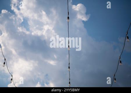 Light bulbs on string wire against beautiful blue cloudy sky background. Long garland of lights. A wedding, event or festival banquet decoration. Stock Photo