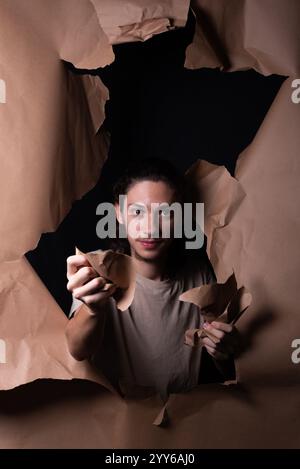 A man coming out through a hole in a sheet of brown paper. Tearing with and showing with his hands the paper. Studio portrait. Stock Photo