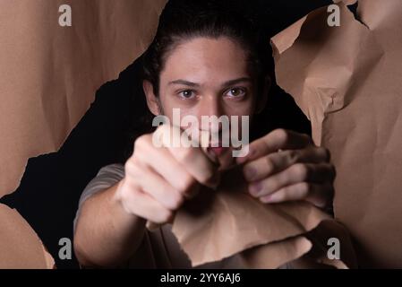 A man coming out through a hole in a sheet of brown paper. Tearing with and showing with his hands the paper. Studio portrait. Stock Photo