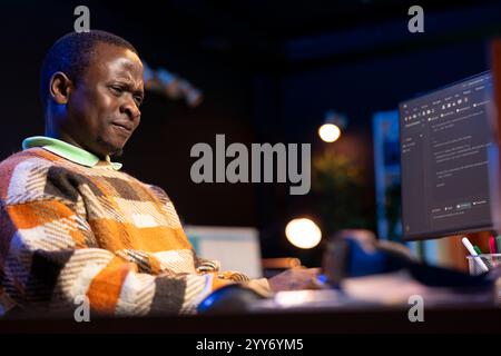 African american self employed developer in home office coding on his computer at workstation. Focused IT specialist doing contracted job from apartment, writing lines of code at desk. Stock Photo