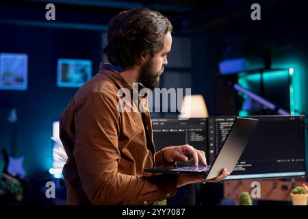 Man doing software quality assurance, reading source code on laptop, crosschecking with computer monitors. IT admin inspecting coding on notebook device, using PC screens at reference point Stock Photo