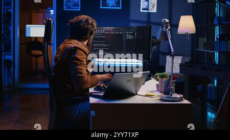 Software programmer putting headphones on while developing code, making time pass faster during remote job shift. IT professional doing multitasking in apartment office while listening music, camera B Stock Photo