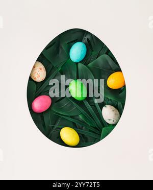 Easter theme featuring an egg cutout filled with colorful Easter eggs and green tropical leaves on white background, symbolizing holiday creativity Stock Photo