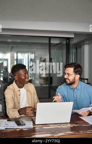 Two busy happy diverse men working on laptops discussing project at work. Stock Photo