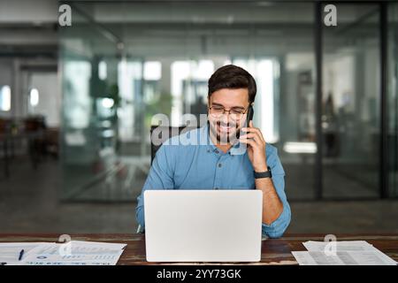 Happy young business man talking on mobile phone using laptop in office. Stock Photo