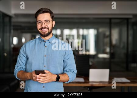 Happy young Latin business man holding phone posing for portrait in office. Stock Photo
