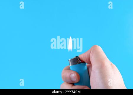 Male hand holding a lighter with a flame on a classic blue background in a studio. Fire and minimalism concept with copy space for text or design Stock Photo