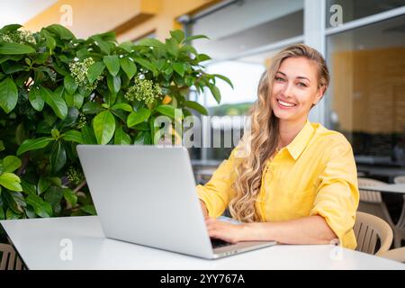 Worldwide work: Happy female developer working on laptop at outdoor restaurant. Smiling confident woman freelancer is looking at camera at cafe. Caucasian lady is working remotely.  Stock Photo