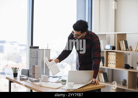 Indian male architect focuses on creating detailed city model for modern urban development, showcasing creativity and precision in office environment with natural light. Stock Photo