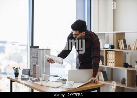Indian male architect focuses on creating detailed city model for modern urban development, showcasing creativity and precision in office environment with natural light. Stock Photo