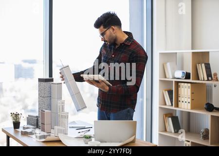 Young Indian male architect works on urban project using digital tools, creating city model in office. Focused on modern design and technology integration. Stock Photo