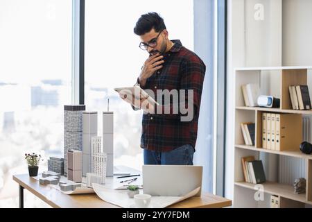 Indian male architect uses digital tablet to design modern city project. He stands in office near building models, displaying concentration and creativity. Stock Photo